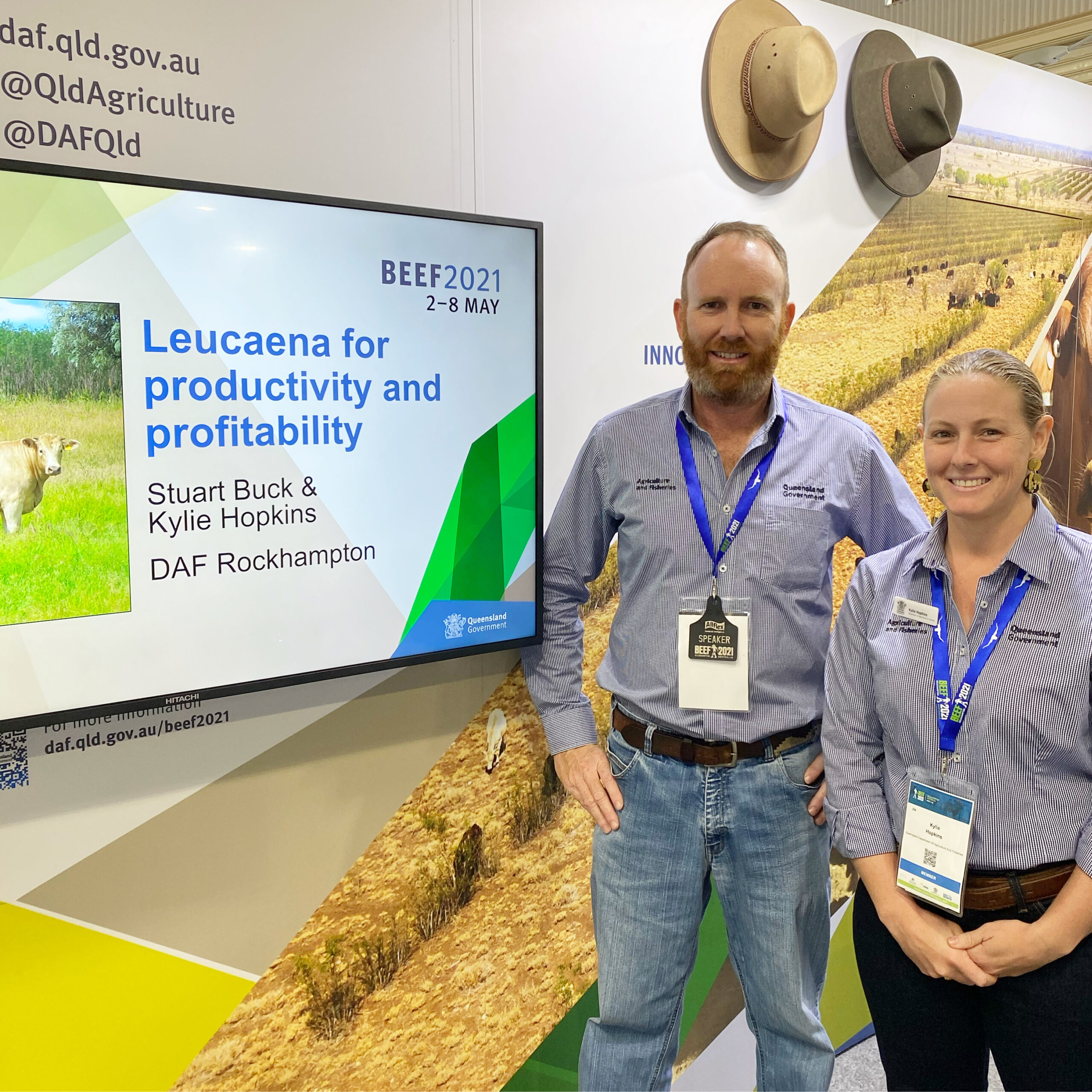 Two DAF staff members standing at the Queensland Government Precinct at Beef Australia 2021. They are standing beside a TV with a presentation about leucaena on it.