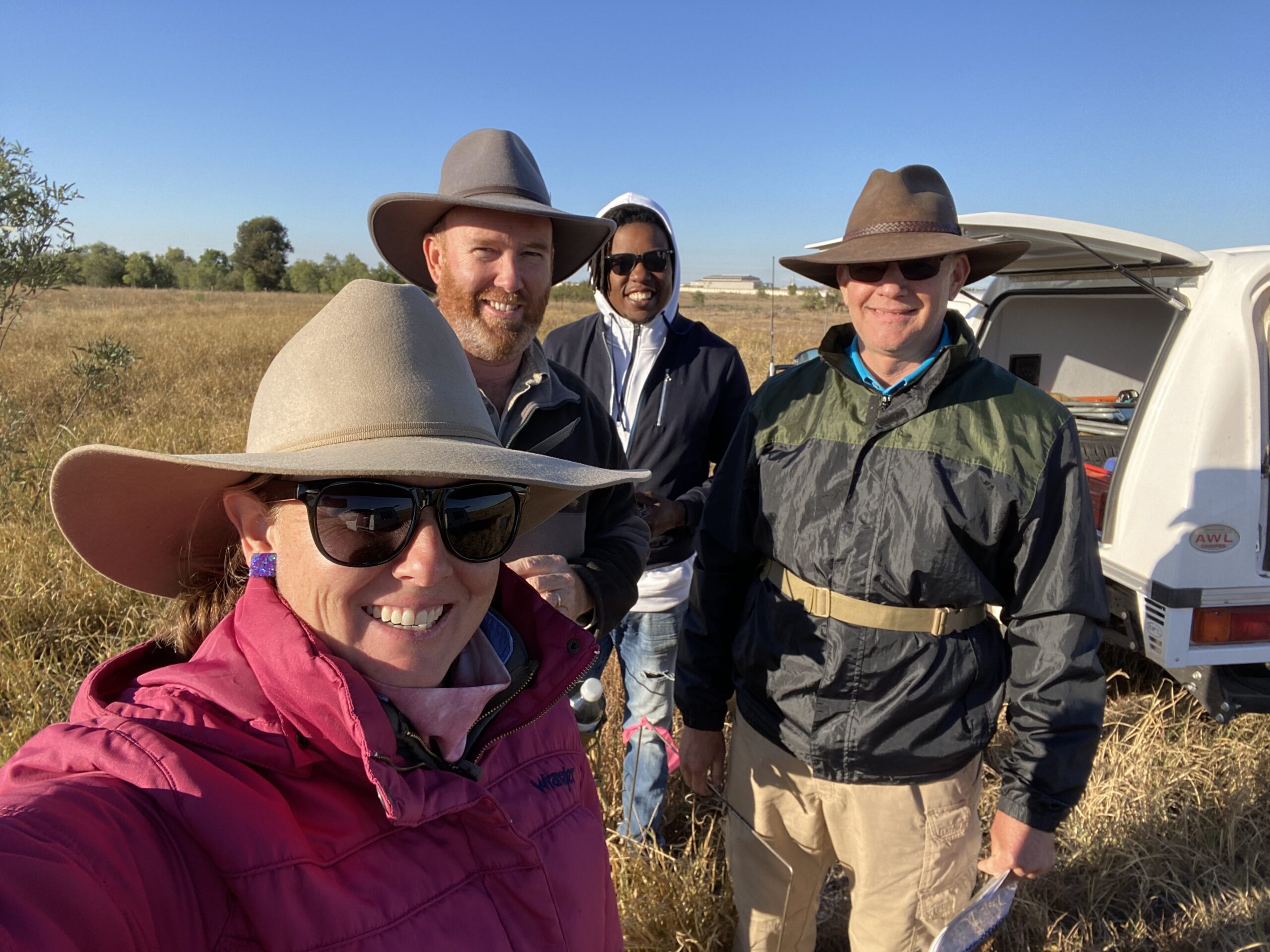 3 men and 1 women taking a selfie in a dry paddock in cold weather, as seen by their large jackets.