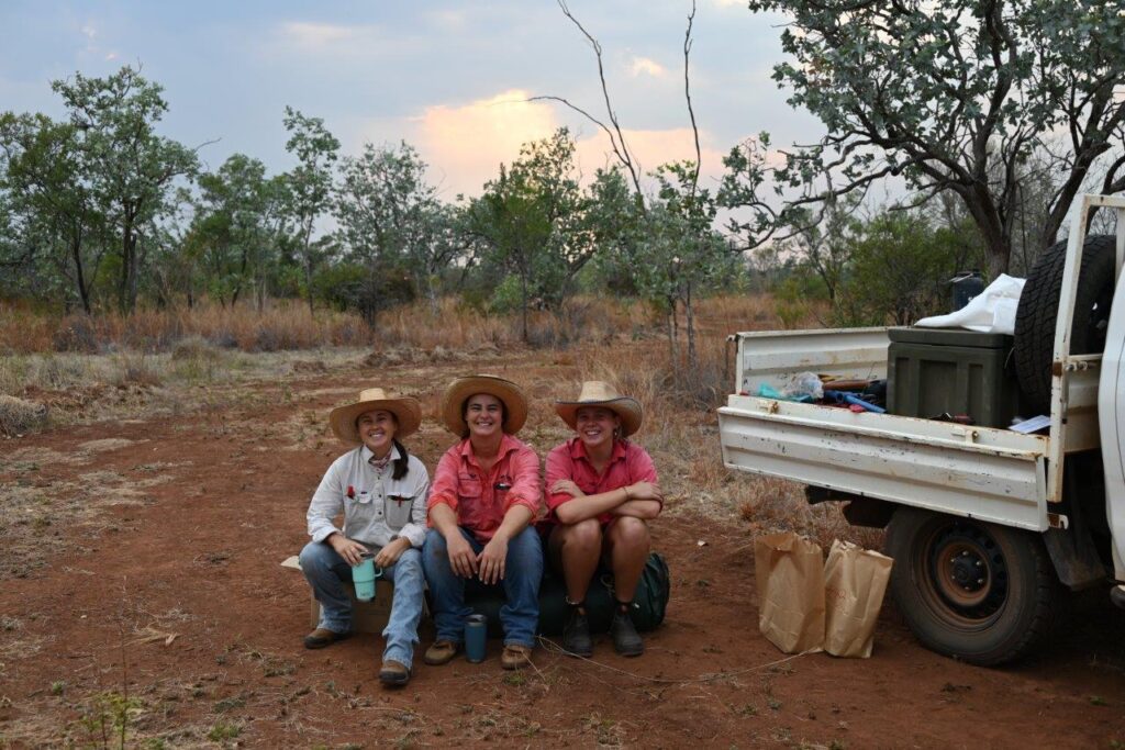 A photo of three young female Kidman Springs Research Station staff sitting beside ute in the paddock having a break from pasture sampling.