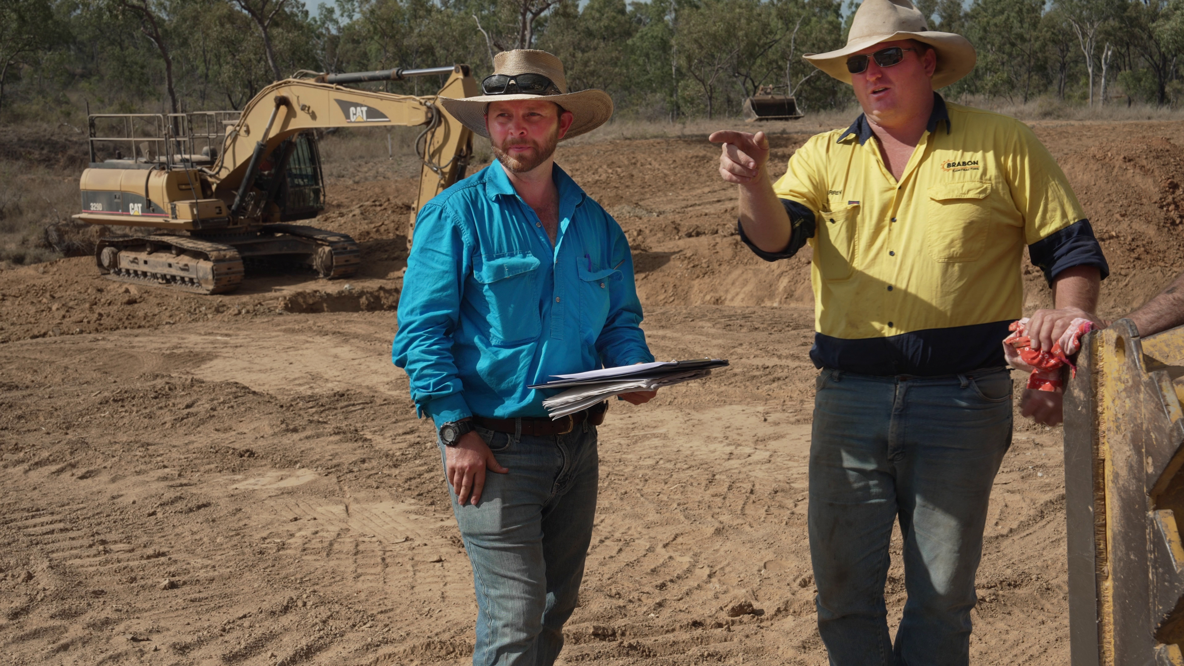 Two men in hats. One (Darren) pointing, the other (Simon) holding a clipboard. Excavator in the background.