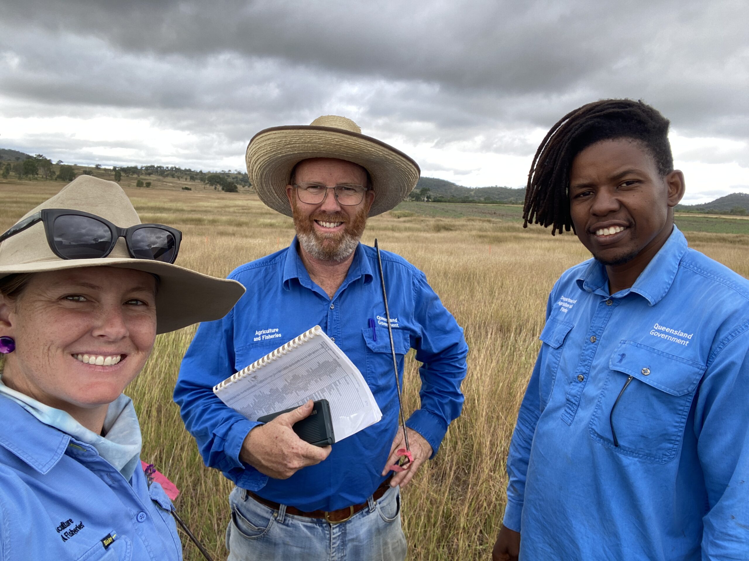 A lady and 2 men standing in a paddock of hayed-off grass holding notebooks and quadrats.