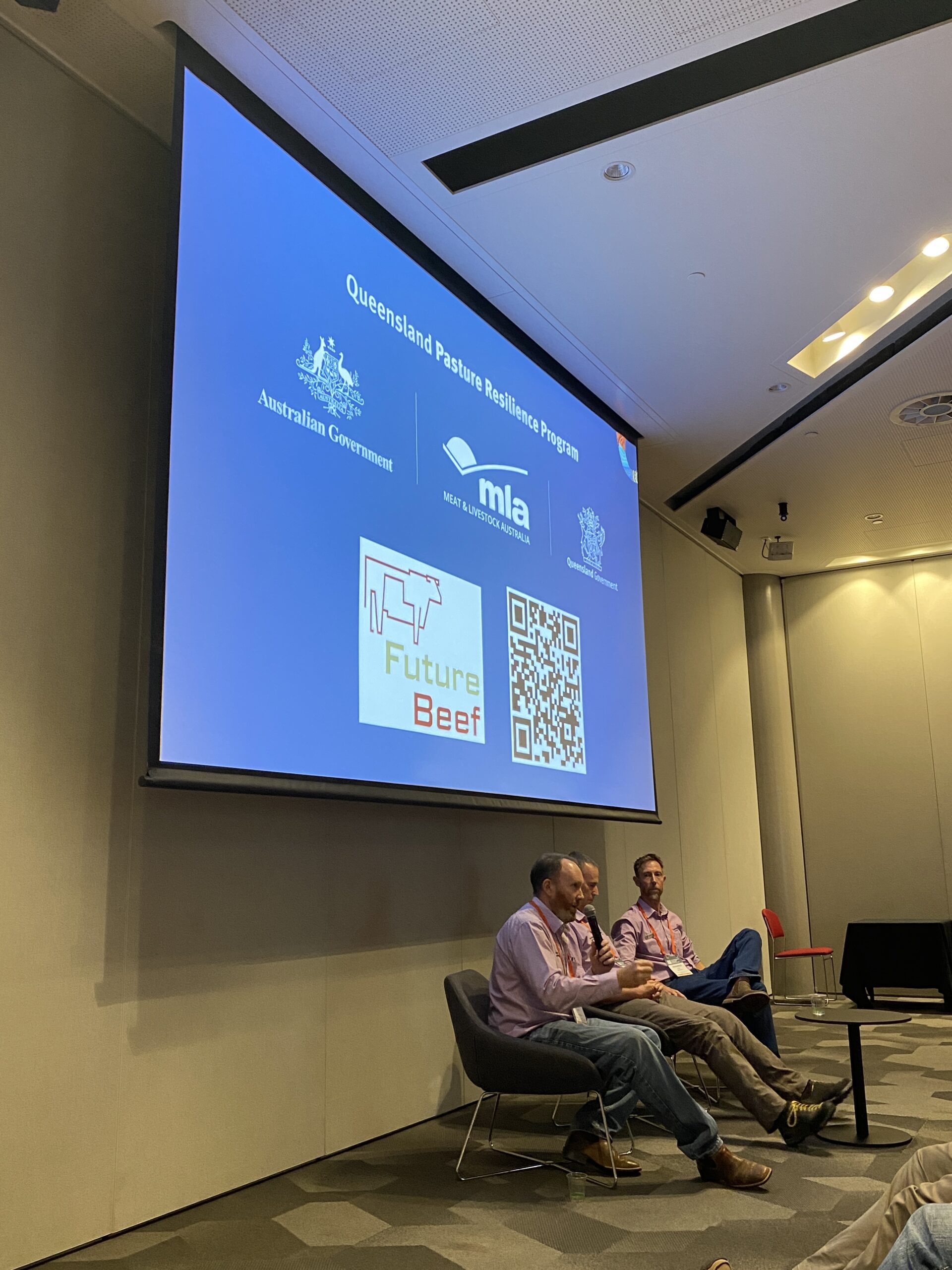 Three men sitting in chairs for a panel session under a large screen promoting the Queensland Pasture Resilience Program.