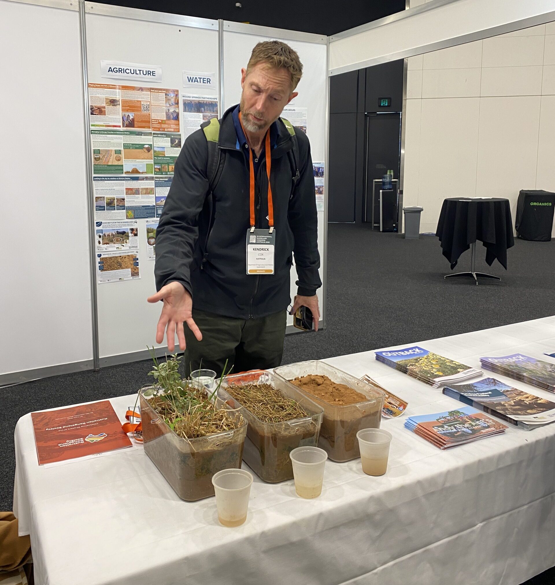 A man pointing at ground cover demonstration made with tubs of soil and vegetation.