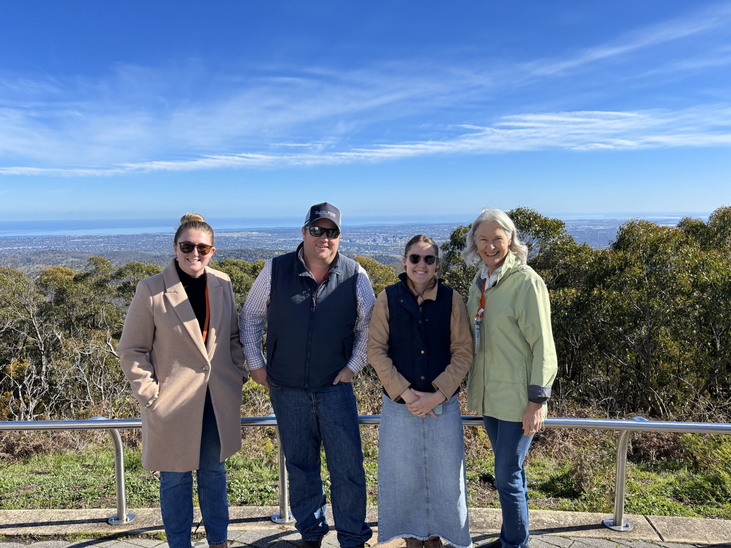 Group of 3 women and 1 man rugged up in the wind at the Mt Lofty summit in Adelaide. The view of the city can be seen in the background.