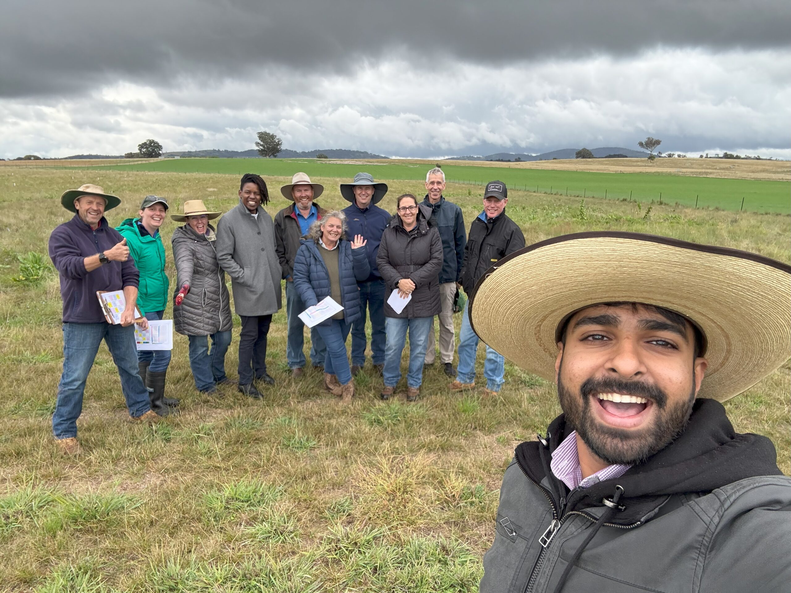 A man with a beard and a big hat taking a selfie in front of a group of people in the paddock. It looks cold and rainy.