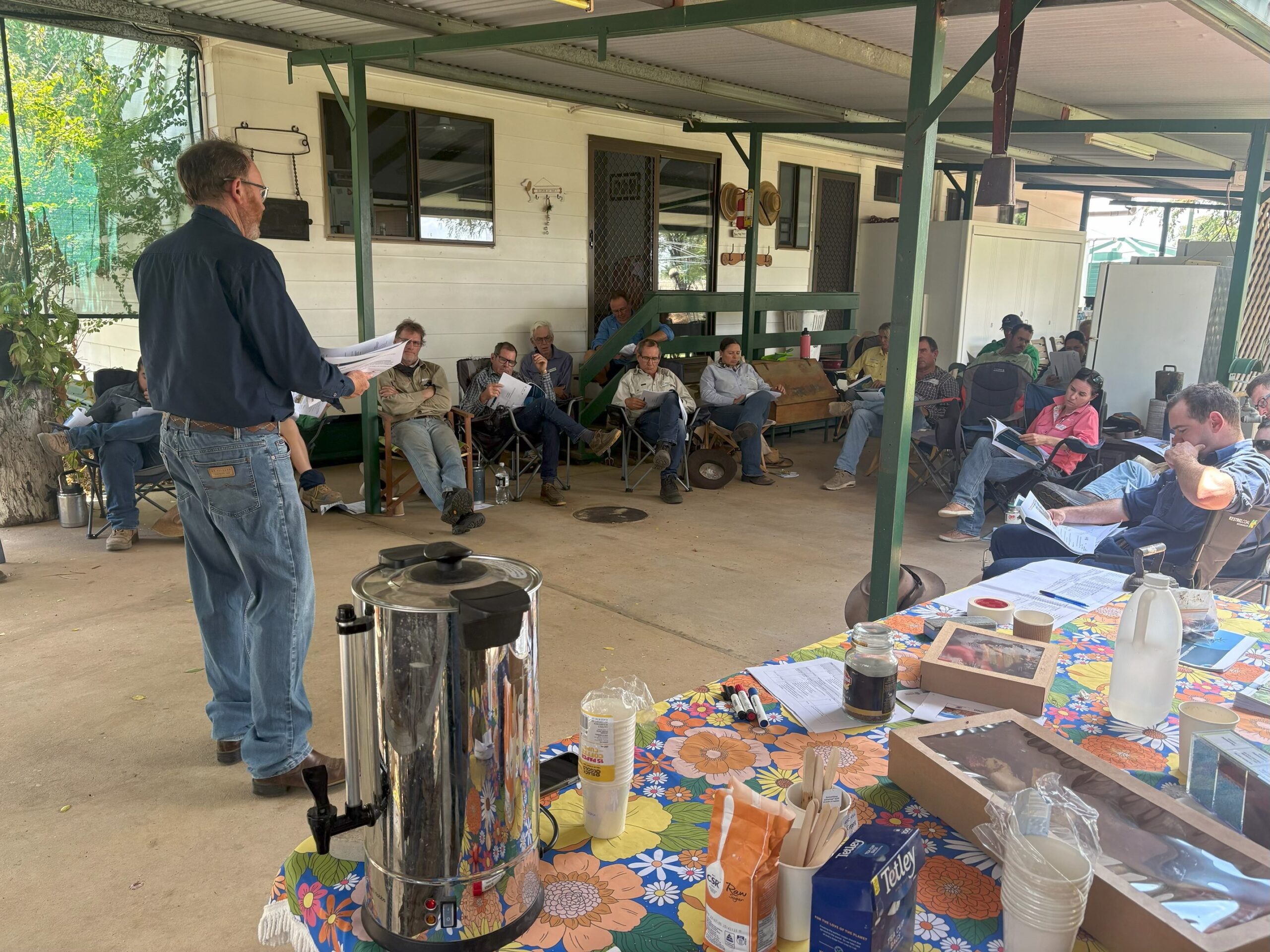 Stuart standing on a verandah with a group of graziers sitting in chairs looks at a booklet.
