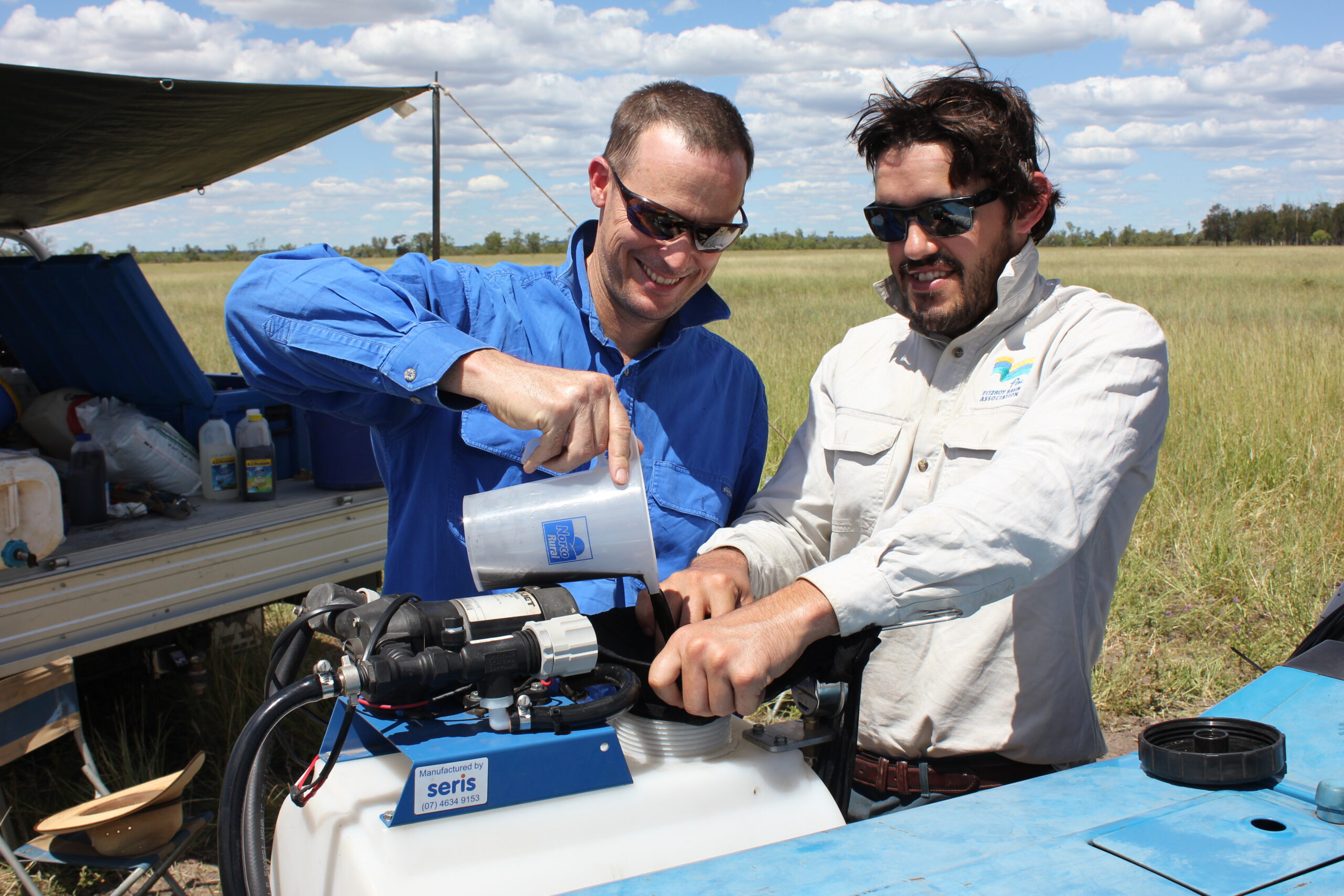 Two men pouring a dark liquid into the top of a spray tank in the paddock.