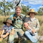 Family photo showing a mum, dad and two kids sitting on a log in a paddock of grass.