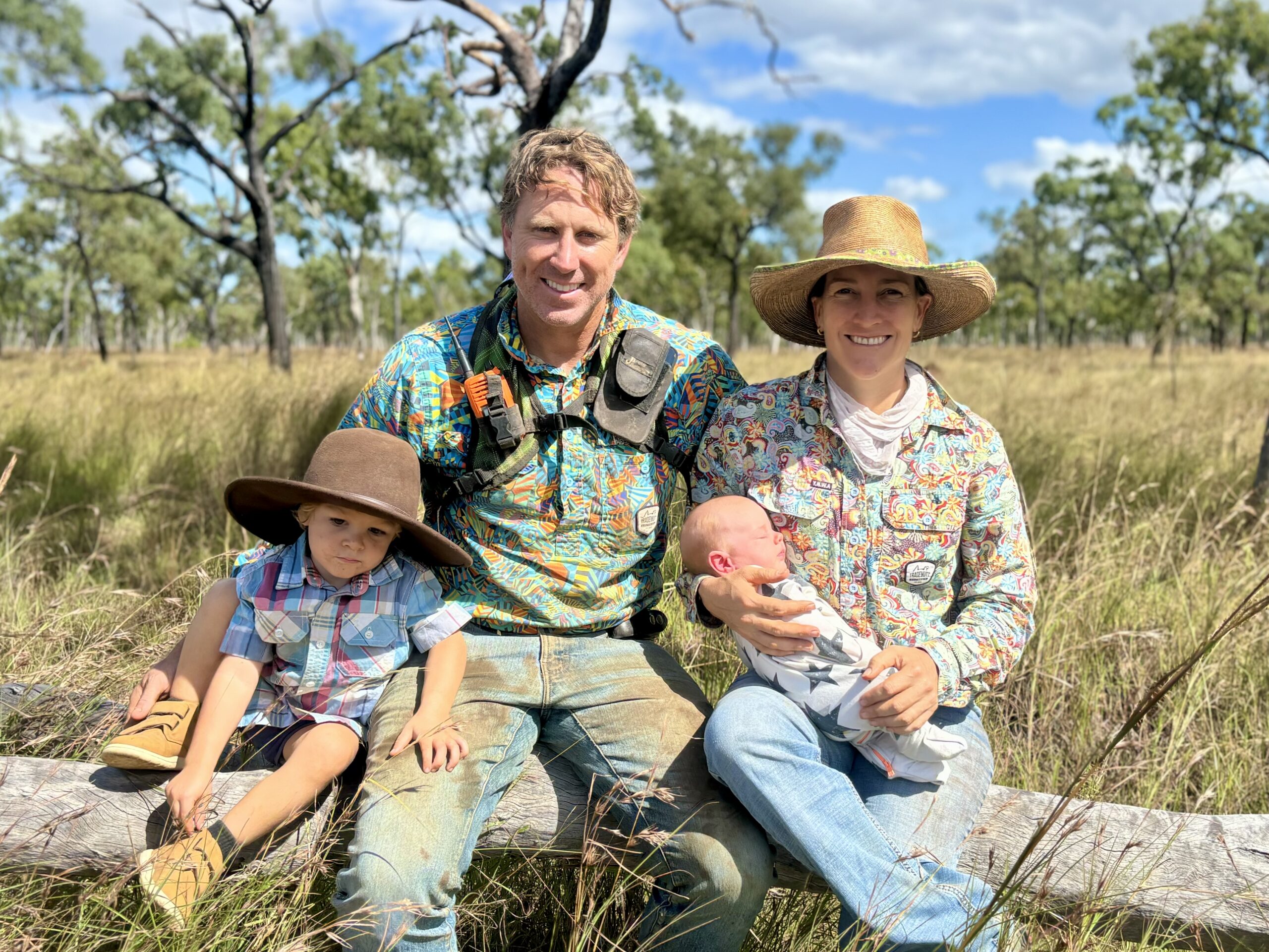 Family photo showing a mum, dad and two kids sitting on a log in a paddock of grass.