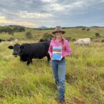 Larissa Zeil-Rolfe, report author, standing in paddock with cows and a copy of the report.
