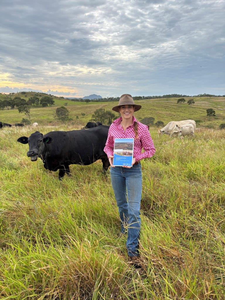 Larissa Zeil-Rolfe, report author, standing in paddock with cows and a copy of the report.